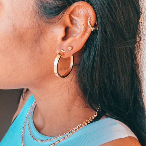 Model wearing layered gold jewelry, necklaces, gold chains, hoop earrings, stud in second earring hole and cartilage piercing